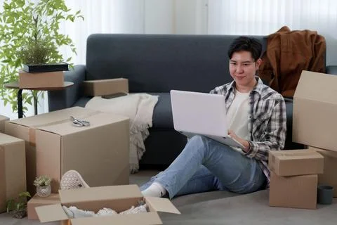 Young Asian man using laptop while sitting on floor surrounded by cardboard.. Stock Photos