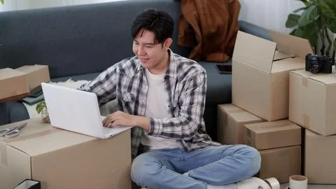 Young Asian man using laptop while sitting among cardboard boxes planning h.. Stock Photos