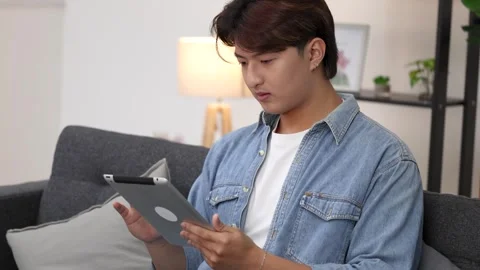Young Asian Man Using Tablet Computer While Sitting on Living Room Sofa Stock Footage 330461923