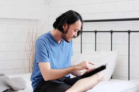 Young asian man using tablet while sitting on bed in white room background, p Foto stock