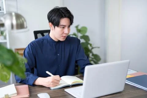 Young Asian man writing notes in journal while working on laptop at bright .. Stock Photos