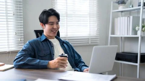 Young Asian professional smiling while working on laptop and holding coffee.. 写真素材