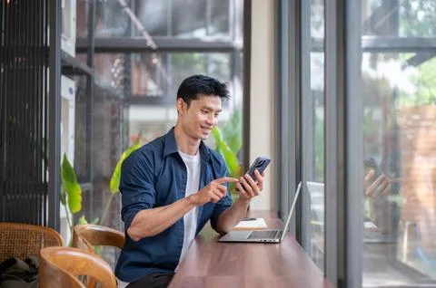 Young Asian professional using smartphone while working on laptop in the cafe Stock Photos