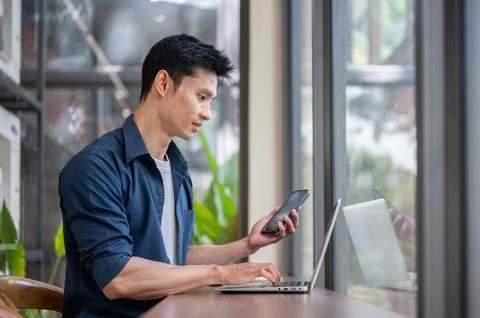 Young Asian professional using smartphone while typing on laptop at cafe tabl Stock Photos