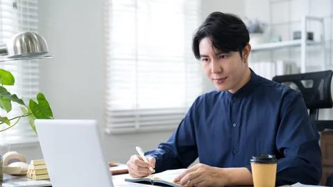 Young Asian professional writing notes in office while working on laptop du.. 스톡 사진