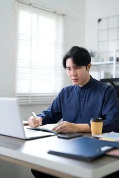 Young Asian professional writing notes in planner while working with laptop.. 스톡 사진