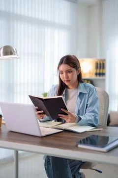 Young Asian student reading notebook while studying with laptop at home des.. 스톡 사진