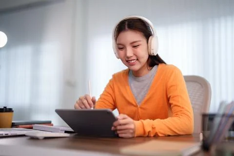 Young Asian student writing notes on digital tablet with headphones during .. Stock Photos