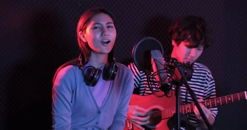 Young asian woman singing while man in background playing acoustic guitar Stockbeeldmateriaal 138932904