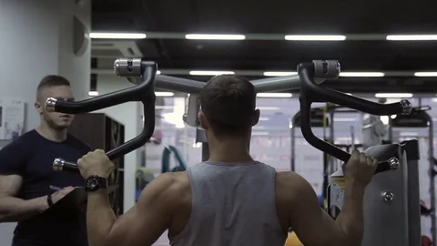 Young athlete is pulling the weight machine in the gym and his male trainer is Video stock 85230935