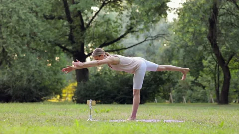 Young athletic female doing yoga online with device and standing in green park Stock Footage
