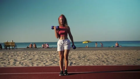 Young athletic girl training with weights on the beach.Dumbbell swings  Stock Footage 198884689