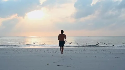 Young athletic guy runs barefoot along the sandy shore towards the ocean Stock Footage 243857046