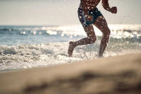 Young athletic man on beach running in water. Focus on lower part of body. .. Stock Photos