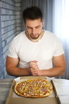 A young athletic man sits at a table and looks at an open package of pizza Stock Photos