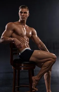 Young athletic shirtless man posing in shorts while sitting on chair in studio Stock Photos