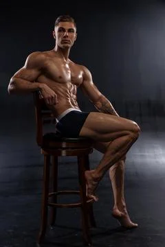 Young athletic shirtless man posing in shorts while sitting on chair in studio Stock Photos