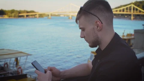 A young attractive bearded guy using his smartphone in a black shirt sits, river Stock Footage 117439250