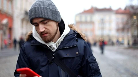 Young attractive brunette man using red smartphone standing outside street. Wint Vidéo 121308873