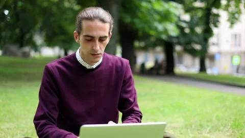 Young attractive brunette man using laptop sitting on grass outside in city. Urb Видео 145756218