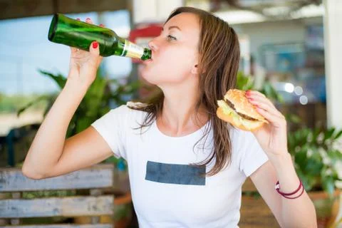 Young attractive girl, with an independent look, drinks beer and eats a burger Stock Photos