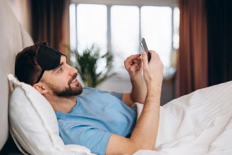 Young attractive man lying in bed checking his messages on his phone in the Stock Photos