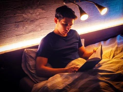 Young attractive man lying in bed and reading a book Stock Photos