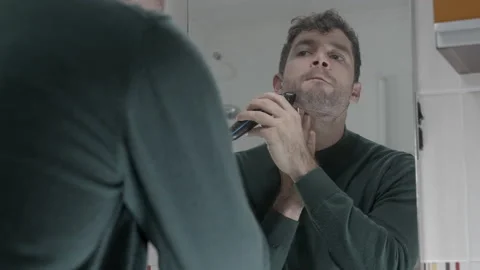 Young attractive man shaving in front of mirror in bathroom electric shaver Vídeos de archivo 230891267