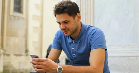 Young attractive man sits and uses the phone on the street old town. Blue t Stock Footage 79208150