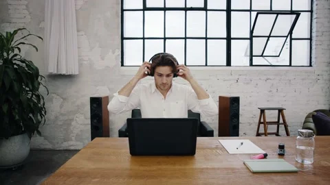 Young attractive man sits in front of his laptop and puts on his headphones Stock Footage 145733748