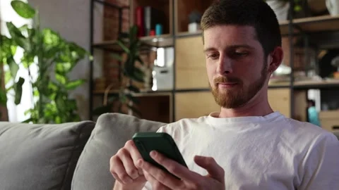 Young attractive man sitting on couch in living room with phone and use phone Video stock 303342468