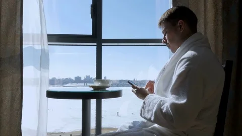 A young, attractive man sitting at a table by the window drinking tea, coffee Video stock 75984536