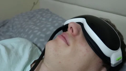 Young attractive man uses eye massager device, lying on the bed Vídeo Stock 104044404