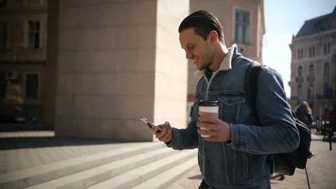 Young attractive man walks down the street holding coffee and using phone Stock Footage
