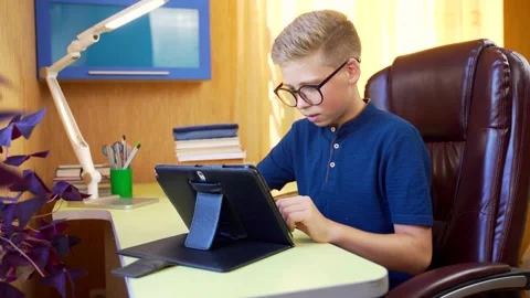 Young attractive schoolboy boy teaches lessons online with tablet at the table.  Stock Footage 138411058