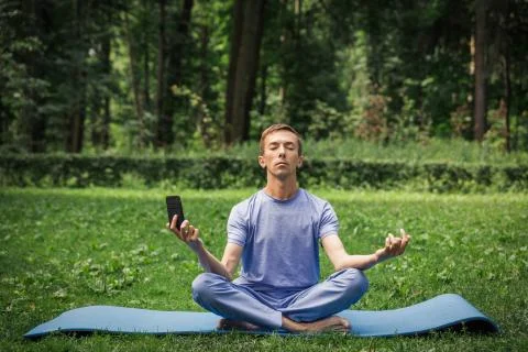 Young attractive white man learns to meditate in the park in front of smartph Stock Photos