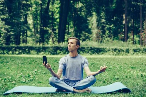Young attractive white man learns to meditate in the park in front of smartph Stock Photos