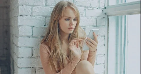 Young attractive woman using mobile phone while sitting near the window at home Stock Footage 82137588
