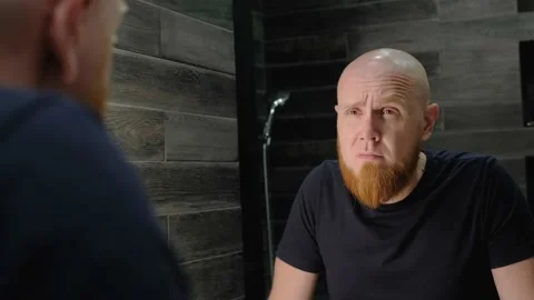 A young, bald guy with a red beard frowns at his reflection in the mirror. Video stock 138998137