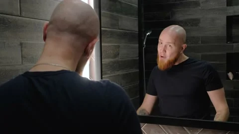 A young bald guy with a red beard points his finger at his reflection in mirror. Stock Footage 138998513