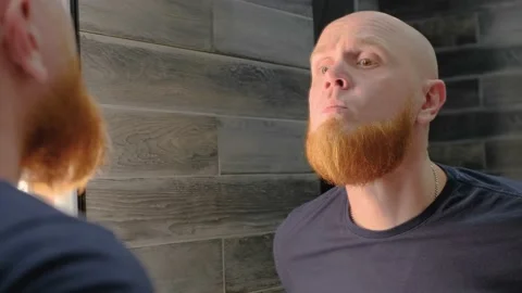 Young bald guy with a red beard appraisingly examines skin of his face and neck Video stock 138999306