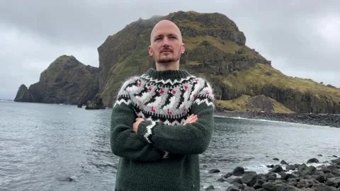 A young bald guy stands against the background in an Icelandic sweater Stock Footage 253110127