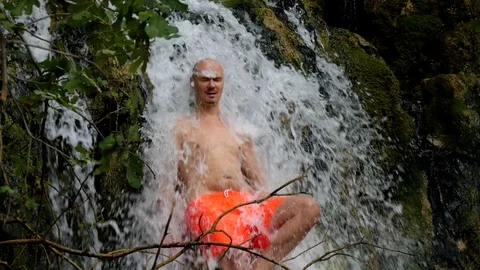 Young bald man in bright orange shorts stands under strong powerful waterfall Stock Footage 122314232