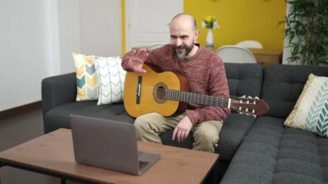 Young bald man having online classical guitar lesson sitting on sofa at home Stock Footage 236535343