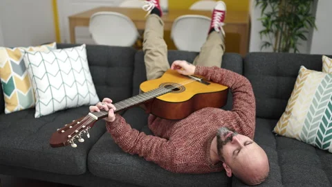 Young bald man playing classical guitar lying on sofa at home Stock Footage 233396991