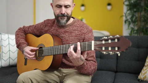Young bald man playing classical guitar sitting on sofa at home Stock Footage 233396997
