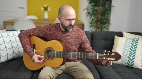 Young bald man playing classical guitar sitting on sofa at home Stock Footage 236535355