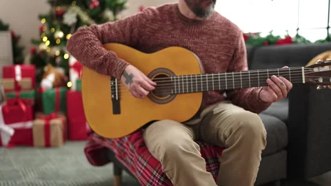 Young bald man playing classical guitar sitting on sofa by christmas tree a.. Stock Footage 255438916