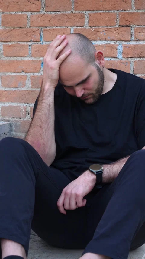 Young bald man with short beard sad sitting against brick wall Stock Footage 295141472