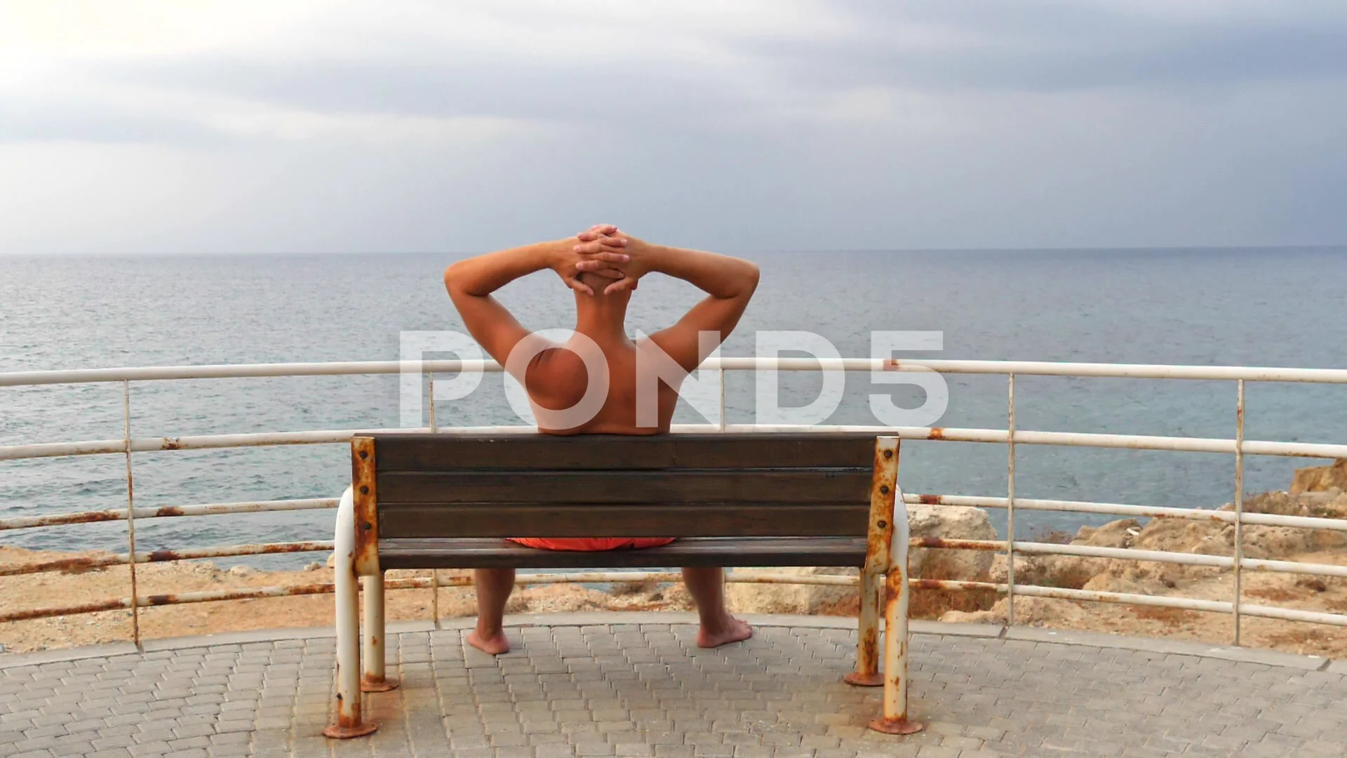 A young bald tanned man with a naked torso sits on an bench with his back